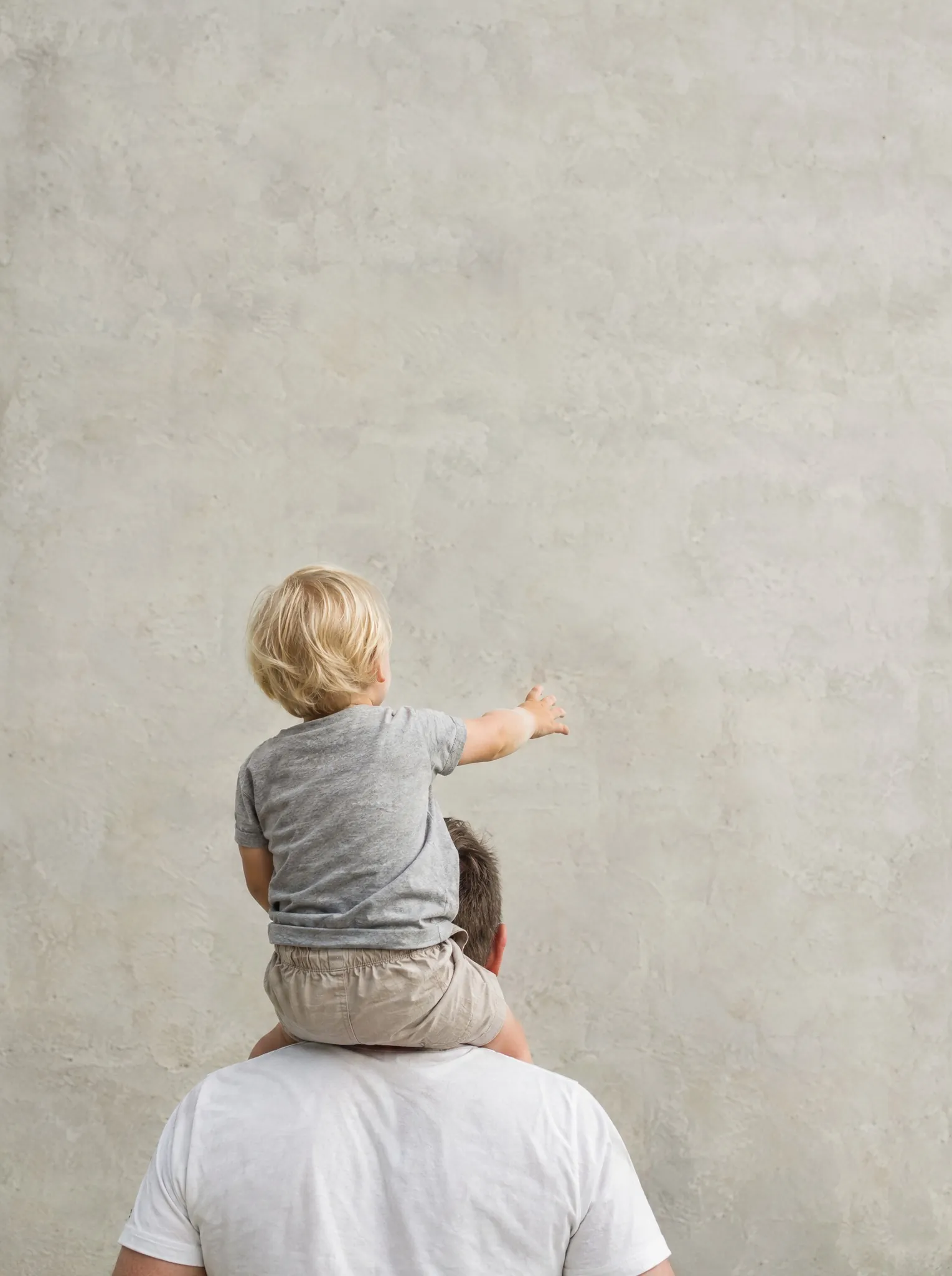 An adult carrying a child on their shoulders by a pale concrete wall, seen from behind